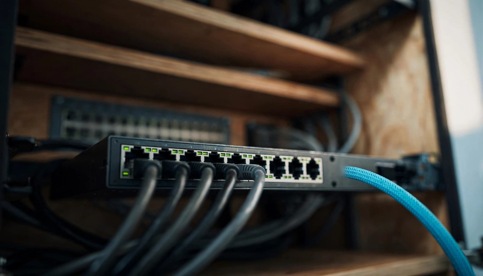 A small Ethernet switch with dark cables and one cyan-sleeved cable on a wood shelf.